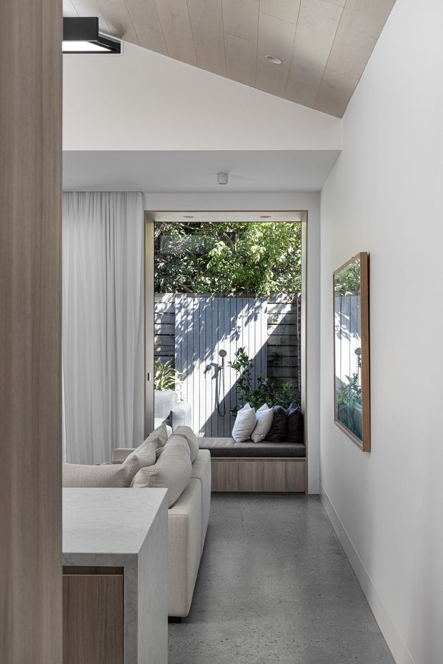 A narrow, bright contemporary living area viewed from an adjoining space. The room features a pale upholstered sofa, built-in window seat with cushions, full-height curtains, a large timber-framed mirror, a light stone or composite counter in the foreground, a polished concrete floor, white walls, and a sloping light timber-panelled ceiling. A glazed opening looks onto a small planted courtyard enclosed by a dark timber fence.