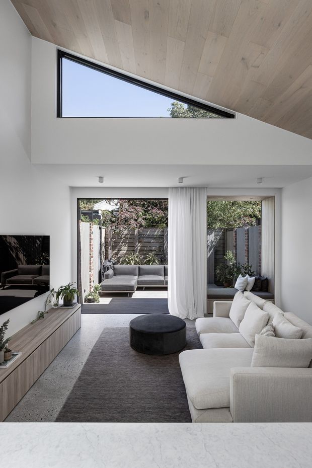 A bright contemporary living room with a large cream sectional sofa, dark round ottoman, grey rug, and timber media joinery. Full-height glazed doors open to a compact courtyard, while a dramatic angled timber-lined ceiling incorporates a long triangular clerestory window. White walls, sheer curtains, and restrained furnishings create a clean, airy interior.
