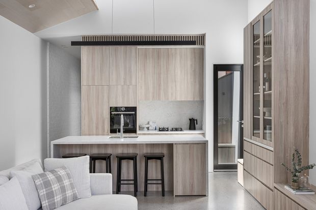 Bright contemporary open-plan kitchen viewed from the living area, featuring full-height light timber cabinetry, a pale stone island with integrated sink, black bar stools, built-in appliances, and a tall glazed display cabinet. A linear ceiling vent and neutral finishes contribute to the streamlined, refined design.