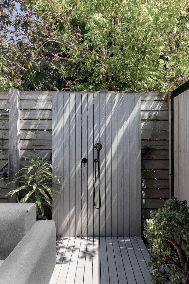 An outdoor shower area enclosed by a timber privacy fence, with vertical grey-painted timber boards forming the shower wall and deck. Matte black shower fittings are mounted centrally, surrounded by leafy planting and a grey outdoor sofa.