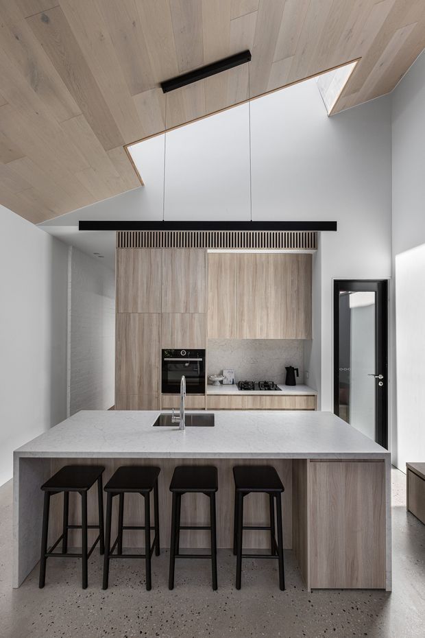 A contemporary kitchen with a large island and four black bar stools. The space features light timber cabinetry and ceiling lining, a pale stone benchtop, integrated appliances, a textured light backsplash, black metal lighting and door frames, and polished concrete-style flooring. A skylight and high ceiling bring in abundant natural light.