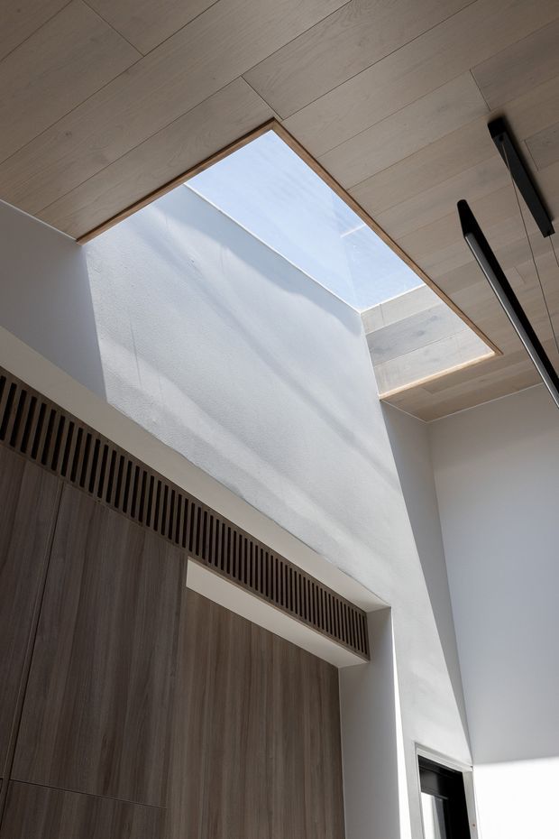 Interior architectural detail showing a tall white plastered void with a large rectangular skylight, pale timber ceiling lining, dark linear pendant lights, and full-height dark timber cabinetry with a slatted ventilation or screening detail above. Strong natural daylight creates a clean, vertical and airy composition.