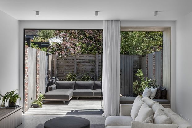 A bright contemporary living room opening onto a compact private courtyard through large glazed doors. The room features a light neutral palette, a large white sectional sofa, dark round ottoman, grey rug, white curtains, recessed ceiling spotlights, and a built-in console. The courtyard includes timber fencing, a paved deck, outdoor seating, and planting.