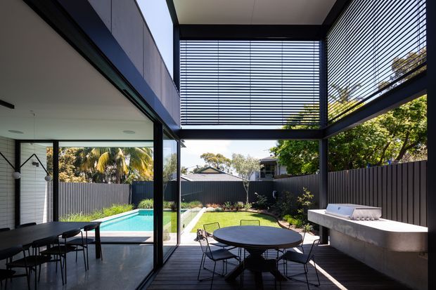 A contemporary covered outdoor courtyard and dining terrace adjoining a glass-walled interior. The space features dark timber decking, a round outdoor dining table with chairs, a built-in concrete outdoor kitchen or barbecue bench, extensive black-framed glazing, and horizontal louvres forming a partially enclosed overhead screen. A lawn and swimming pool are visible beyond, with a high dark boundary fence and established planting.