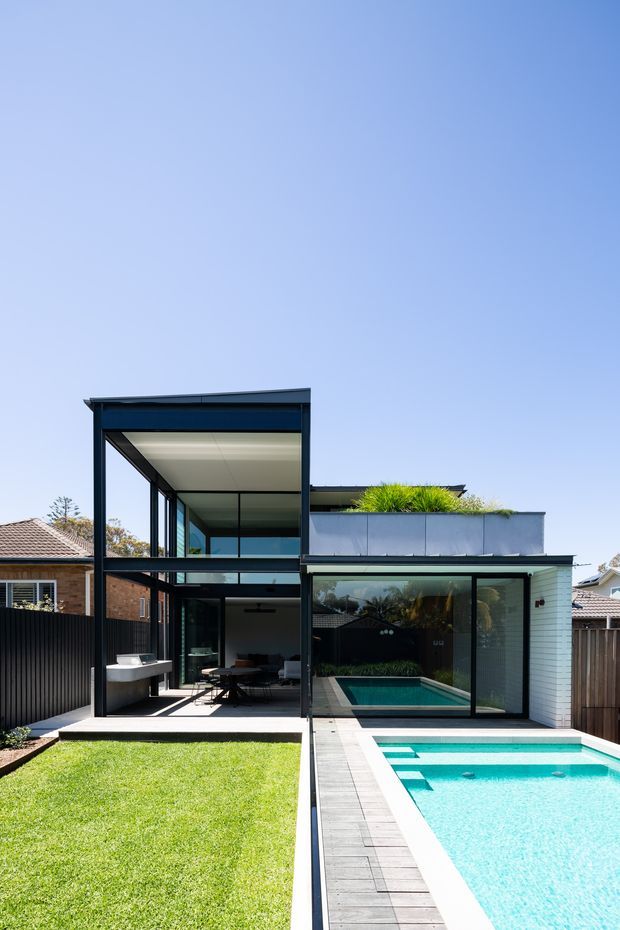Contemporary two-storey residential extension viewed from the backyard, with extensive black-framed glazing, a covered outdoor dining area, rooftop planting, and a bright rectangular swimming pool bordered by timber decking and lawn. Neighbouring suburban homes and fences are visible around the property under a clear blue sky.