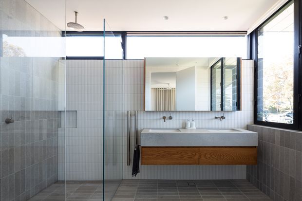 Bright contemporary bathroom with a floating double vanity, timber drawer cabinetry, wall-mounted basins and taps, large rectangular mirror, glass shower enclosure, and floor-to-ceiling windows. The room uses small-format vertical white wall tiles, dark grey floor and lower wall tiles, black window framing, and clean minimalist detailing.