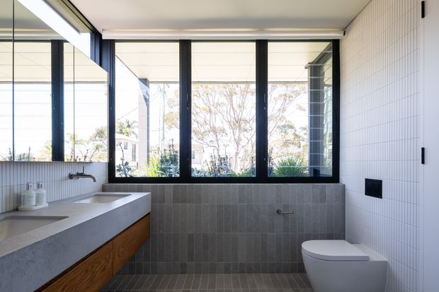 Bright contemporary bathroom with a floating timber vanity and pale concrete-look benchtop, wall-mounted basin, freestanding bathtub partially visible at left, wall-hung toilet, and large black-framed windows. White small-format tiled walls contrast with darker grey vertical wall and floor tiles, creating a clean, modern interior with abundant natural light.