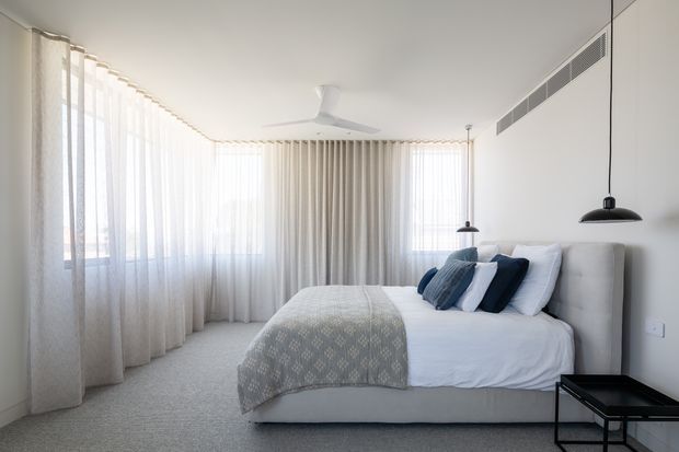 Bright, serene contemporary bedroom with a large upholstered bed, layered white and blue bedding, full-height sheer curtains wrapping around corner windows, pale carpeting, minimalist pendant lights, and a white ceiling fan. A linear air-conditioning vent runs along the upper wall, while a small black side table adds contrast.