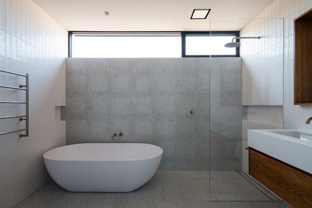 A contemporary bathroom featuring a freestanding white bathtub, large grey stone-look tiled wall and floor surfaces, a frameless glass shower enclosure, white vertically patterned wall tiles, and a floating timber vanity with a white basin. High horizontal glazing provides natural light, while a wall-mounted towel rail and minimal fittings reinforce the clean, refined design.