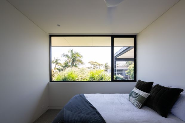 A compact, contemporary bedroom with a neatly made bed positioned beneath a large black-framed horizontal window. The room has plain white walls and ceiling, a neutral carpeted floor, dark bedding and pillows, and bright natural light overlooking lush tropical vegetation and a neighbouring modern structure. The interior is simple and uncluttered.