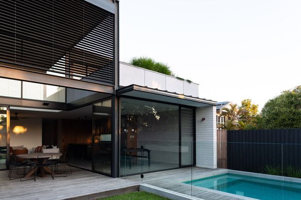 Modern two-storey residence with a covered outdoor dining and living area opening onto a compact rectangular swimming pool. The house features extensive black-framed glazing, horizontal timber-like louvres forming a sunshade, pale masonry walls, a timber deck, and a dark privacy fence. The image is photographed from the backyard and emphasizes the indoor-outdoor connection and pool terrace.
