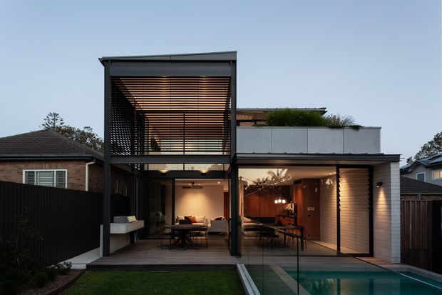 Contemporary two-storey residential extension viewed from the backyard at dusk, with a covered outdoor dining and living zone opening onto a swimming pool. The facade combines dark slender framing, extensive glazing, horizontal timber screening and a rooftop planter terrace, with warm interior lighting visible inside.