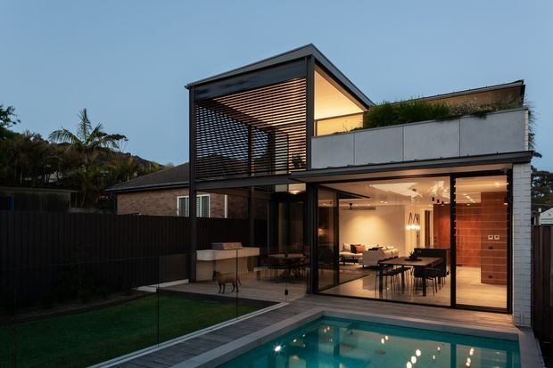 Contemporary multi-level residential house viewed from the rear pool area at dusk. The design features extensive floor-to-ceiling glazing, a dark metal and timber-look slatted upper pavilion, a flat-roofed planted terrace, brick walls, and an illuminated open-plan dining and living area overlooking the swimming pool. A fenced lawn and landscaped surroundings frame the outdoor entertaining space.