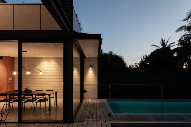 Dusk exterior view of a contemporary single-storey residence opening onto a timber deck and swimming pool. The visible dining area has full-height glazing, warm pendant lighting, a timber-toned horizontal wall finish, and dark-framed doors. The pool and deck occupy the foreground, with a dark privacy fence and tropical planting beyond.