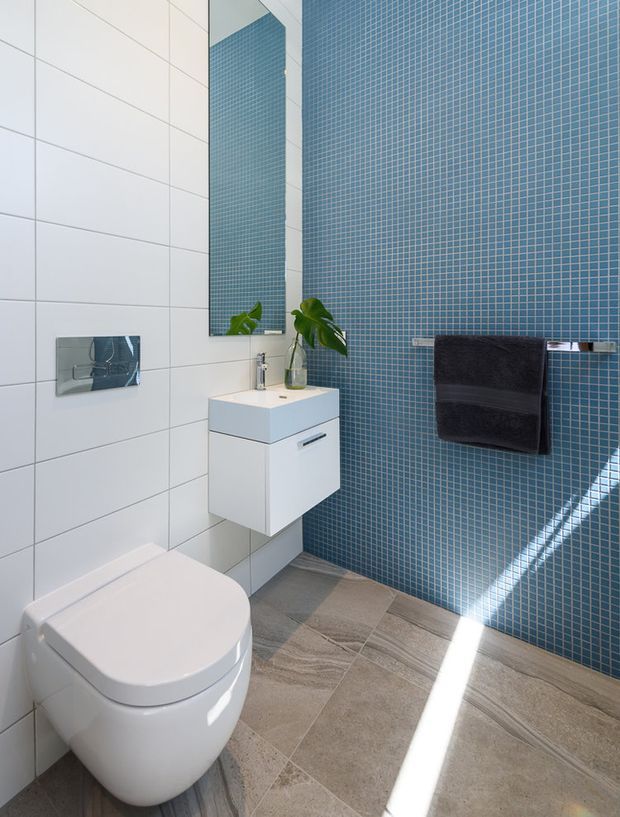 Bright contemporary bathroom with a wall-hung white toilet and floating vanity, large white wall tiles, a blue mosaic-tile feature wall, grey-beige stone-look floor tiles, a frameless mirror, chrome flush plate and tapware, and a dark towel rail. Sunlight creates a strong diagonal highlight across the floor.