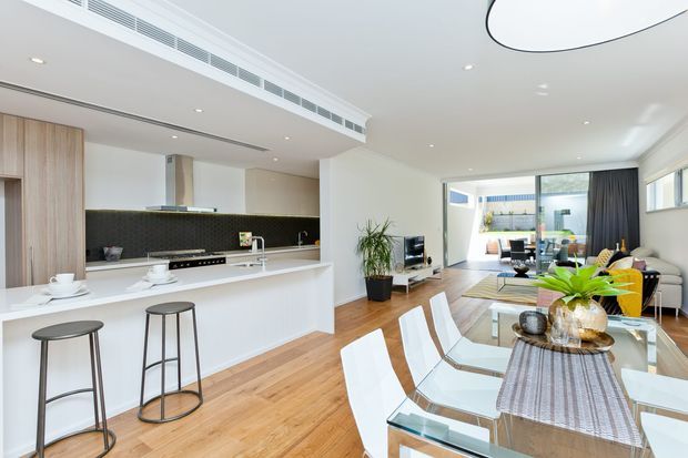 Bright open-plan contemporary interior combining a white kitchen with island seating, a dining area with a glass table and white chairs, and a living area beyond. Warm timber flooring, light timber cabinetry, a dark tiled splashback, expansive glazing, and minimalist furnishings create an airy, polished space.