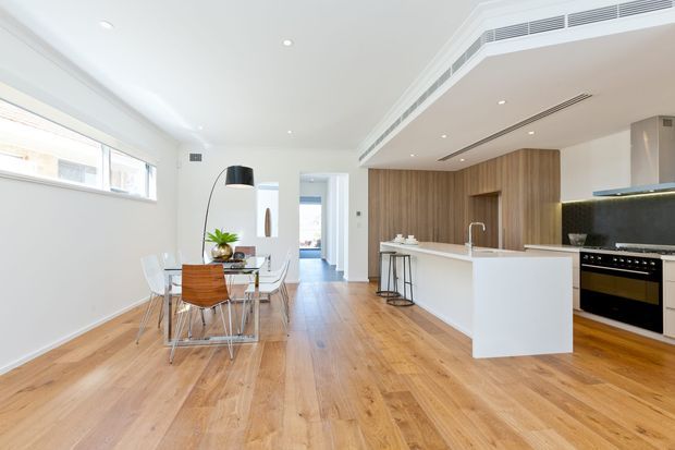 Bright open-plan kitchen and dining area with a white island, timber cabinetry, integrated appliances, and a timber floor. A glass dining table with modern chairs sits beside the kitchen, while concealed air-conditioning grilles and recessed lighting contribute to a clean contemporary fit-out.
