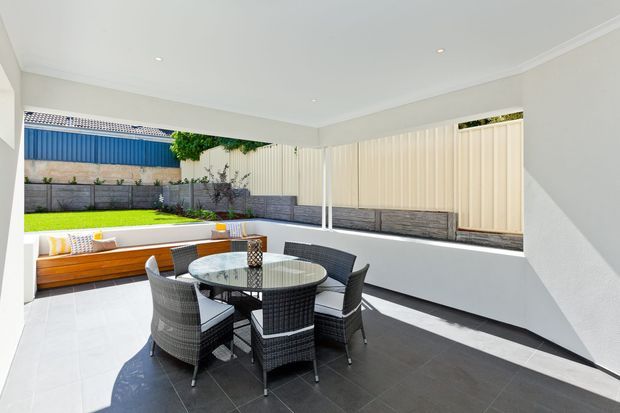 Bright covered outdoor entertaining area with a round glass dining table and woven chairs, adjoining a built-in timber bench seat and landscaped courtyard. The space has pale tiled walls and ceiling, dark floor tiles, white structural columns, and large open sides with privacy screening and garden views.