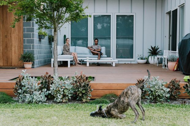 A contemporary outdoor living deck adjoining a light-coloured home, with built-in bench seating, a pale outdoor sofa, potted plants, timber decking, and large glazed doors. A rendered block wall, timber entry door, landscaped planting, and a dog in the foreground are also visible.