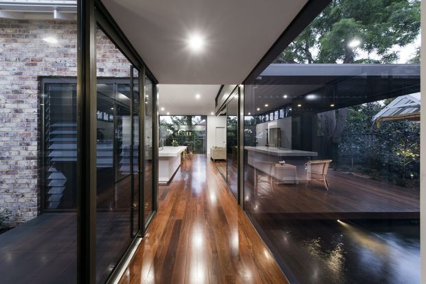 A contemporary open-plan interior corridor with polished dark timber flooring, extensive black-framed glazing, and views into a modern kitchen and dining area. The glazed walls connect the interior to a covered timber deck and landscaped courtyard, creating a strong indoor-outdoor relationship. Exposed brickwork is visible on the left, while recessed ceiling lights provide an even, warm illumination.