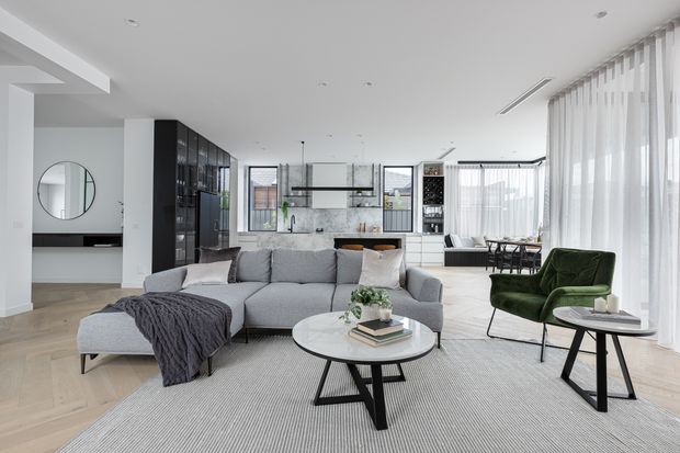A bright, open-plan contemporary living room with a large grey sectional sofa, green accent armchair, round coffee tables, and a textured area rug. The space opens directly to a modern kitchen and dining area, with extensive sheer floor-to-ceiling curtains, pale timber flooring, white walls, black-framed glazing, and integrated dark cabinetry.