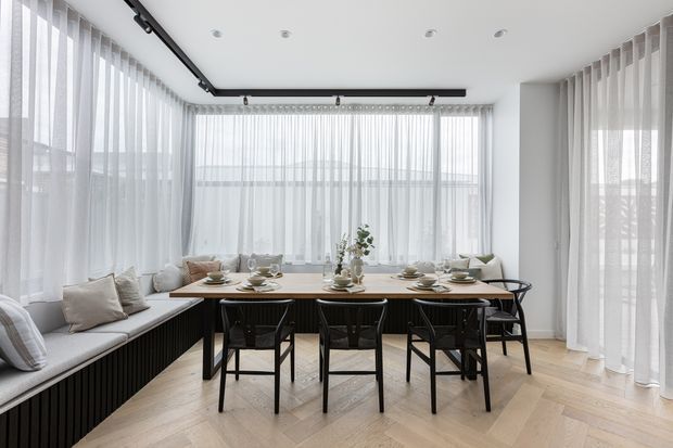 A bright, contemporary dining area with a long timber dining table surrounded by black chairs, set within a glazed corner enclosure. A built-in window bench with light cushions runs along the perimeter, while sheer white curtains soften the abundant daylight. The pale herringbone timber floor, white walls, and black ceiling-mounted track lighting create a clean, refined interior.