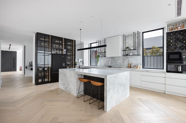 A bright, spacious contemporary kitchen with a large marble-look island and integrated seating, white flat-panel cabinetry, a matching splashback, black-framed glazing, and a dramatic black glass-fronted pantry or display cabinet. The kitchen opens toward a hallway and appears carefully styled with warm timber herringbone flooring, dark metal details, open shelving, and integrated appliances.