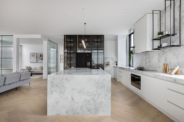 Bright open-plan contemporary kitchen with a large marble-look island, white flat-panel cabinetry, integrated appliances, black-framed glazed partitioning, and open black metal shelving. A living area is visible beyond the kitchen, and the space features light timber herringbone flooring and a clean neutral palette.