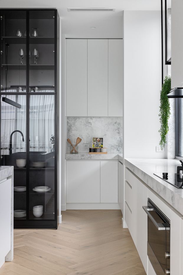 Bright, narrow contemporary kitchen with white handleless cabinetry, pale marble-look benchtops and splashback, integrated appliances, and light herringbone timber flooring. A tall black-framed glass display cabinet with crockery and glassware provides contrast, while a trailing plant and large windows add softness and natural light.