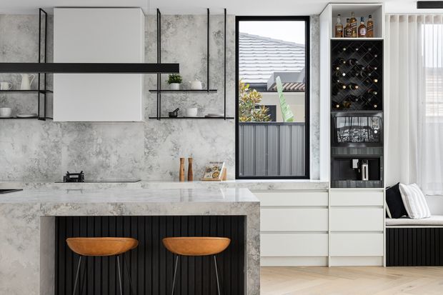 Contemporary kitchen with a concrete-look feature wall, pale stone benchtops, white cabinetry, open black metal shelving, and a dark fluted island base with two curved timber stools. A built-in wine rack and drinks storage sit beside the window, while a window seat and sheer curtains add softness. The room has a refined monochromatic palette with warm wood accents and strong vertical detailing.