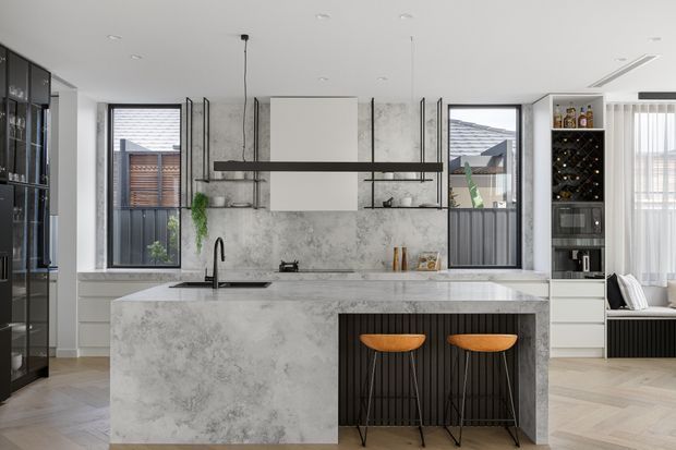 A refined contemporary kitchen centred on a large marble-look island with an integrated black sink and two timber-seated bar stools. The space features dark fluted cabinetry, tall black-framed glazing, a full-height marble-look splashback, integrated appliances, a wine display, and a linear black pendant light. Herringbone timber flooring and abundant natural light add warmth and contrast.