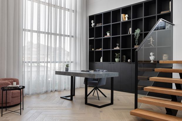 A refined contemporary interior featuring a compact home office beside an open timber staircase. The room has a black built-in shelving and cabinetry wall, a dark stone-look desk with a black metal base, pale herringbone timber flooring, sheer floor-to-ceiling curtains, and a blush armchair. The staircase has floating timber treads, a dark structural frame, and a glass balustrade.