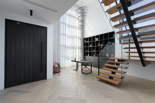 A bright, high-ceilinged contemporary entry and living space featuring a dramatic open-tread timber and black metal staircase with glass balustrades. Pale herringbone timber flooring, a tall sheer-curtained window, black built-in shelving, a dark vertical-panel entry door, a console table, and sculptural ring pendant lights create a refined minimalist interior.