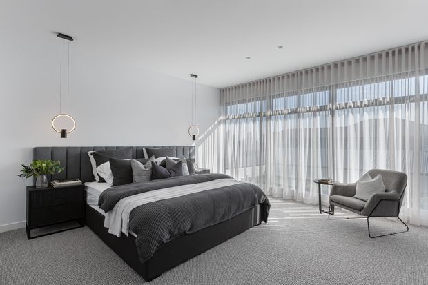 A bright, spacious contemporary bedroom with a large dark upholstered bed, layered grey bedding and cushions, a black bedside cabinet, slim pendant lights, and a grey lounge chair beside full-height sheer curtains. Floor-to-ceiling glazing brings in abundant natural light, creating a refined monochromatic interior.
