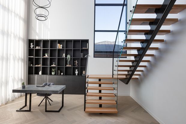 Bright contemporary interior featuring a floating open-riser staircase with timber treads, black steel structure, and clear glass balustrade. A double-height-feeling space includes a dark built-in display wall, a minimalist dining or work table, tall sheer-curtained glazing, and a sculptural ring pendant light. Light herringbone timber flooring and neutral white walls create an airy, refined setting.