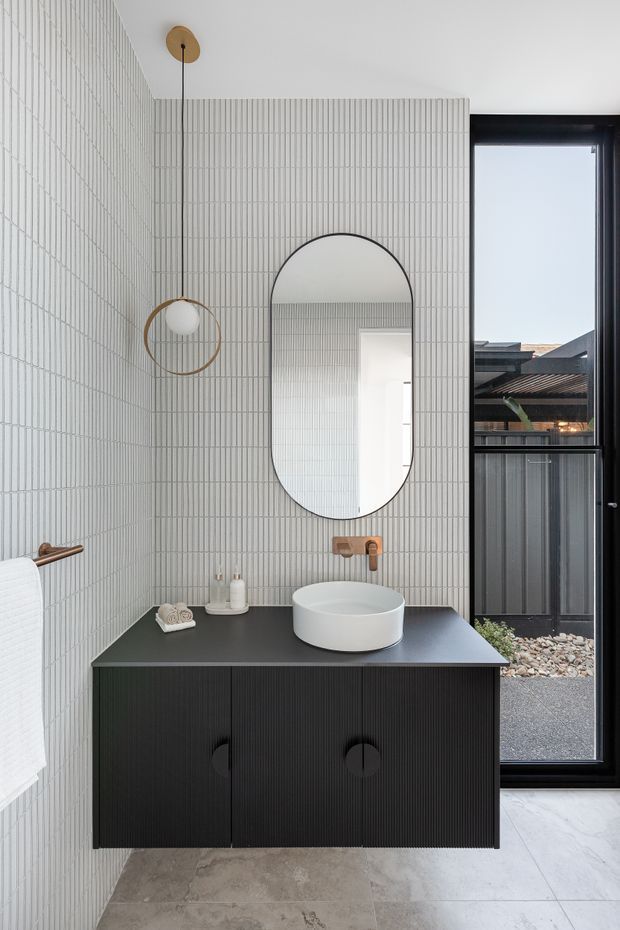 A contemporary bathroom vanity area with vertically ribbed light wall tiles, a floating dark fluted vanity with integrated storage, a white vessel basin, brushed brass tapware and wall-mounted circular pendant. An arched mirror hangs above the basin, while a large black-framed glazed door or window provides exterior light. The floor is finished in large pale stone-look tiles.