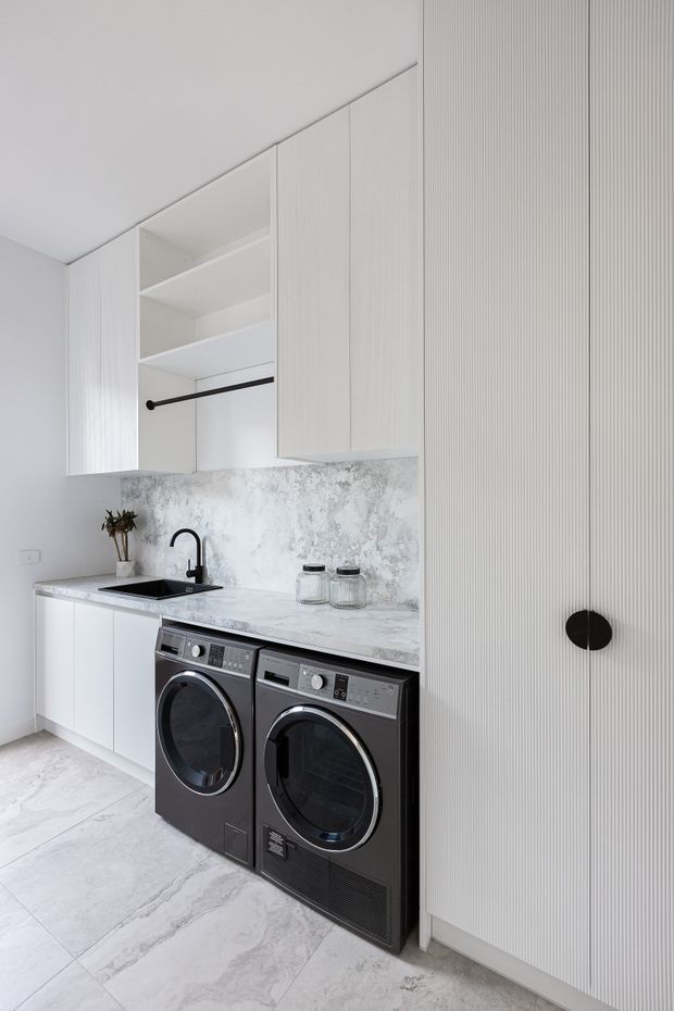 Bright, compact contemporary laundry with white vertically fluted cabinetry, open shelving, a pale marble-look benchtop and matching textured splashback. Black tapware, sink, hanging rail and front-loading washer and dryer provide contrast, while large light stone-look floor tiles continue the restrained monochromatic scheme.