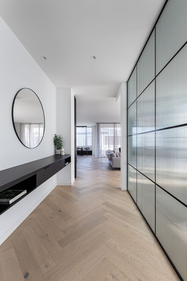 A bright, minimalist residential hallway with a pale timber herringbone floor, white walls and ceiling, and a long dark built-in console beneath a large round mirror. A full-height black-framed ribbed glass partition lines the right side, while the hallway opens toward a living area with soft furnishings and sheer curtains.