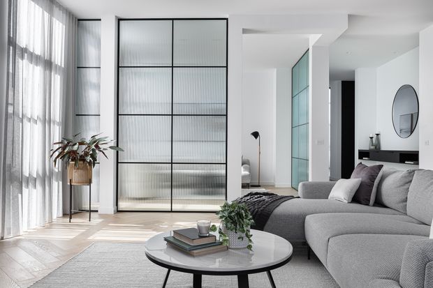 Bright contemporary living room with a large light-grey sectional sofa, round coffee table, tall sheer-curtained windows, pale timber flooring, and a black-framed reeded-glass partition door. A plant, layered books, and neutral furnishings add warmth, while a hallway and reflective glass panels extend the open-plan interior.