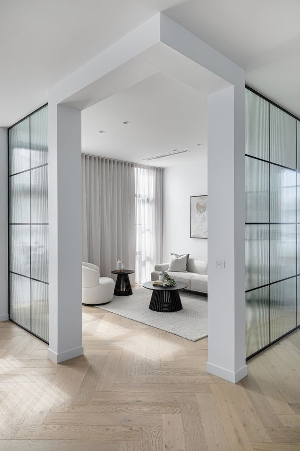 A bright, refined living room viewed through a wide white architectural opening. The space features pale herringbone timber flooring, black-framed fluted glass screens on both sides, soft grey curtains, a light sofa and rounded armchair, black sculptural coffee tables, and neutral artwork. Recessed ceiling lighting and abundant filtered daylight create an airy contemporary interior.