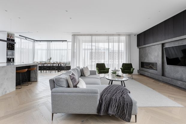 Open-plan contemporary living area with a large light-grey sectional sofa facing a dark feature media wall and fireplace. The space includes a refined kitchen with stone island, dining area, floor-to-ceiling sheer curtains, pale herringbone timber flooring, green lounge chairs, and a neutral layered interior palette.