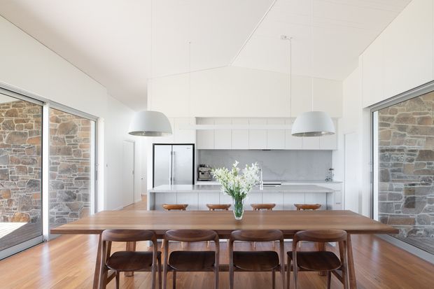 Bright open-plan dining and kitchen interior with a long solid timber dining table and matching wooden chairs in the foreground. A white kitchen with island, integrated cabinetry, refrigerator and marble-look splashback occupies the rear, while large glazed openings on both sides reveal exposed stone walls. White pendant lights and a white paneled ceiling create an airy, refined setting.