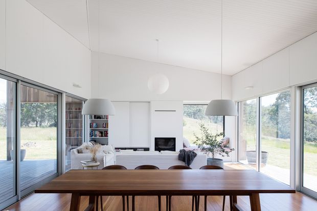 Bright open-plan dining and living area with a long dark timber dining table in the foreground, surrounded by large glazed sliding doors and windows overlooking a leafy landscape. The room has white walls and ceiling, integrated white cabinetry, contemporary pendant lights, a white sofa, shelving, and a central fireplace. The space feels airy, calm, and connected to the outdoors.