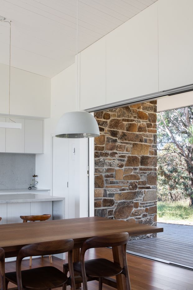 A bright, restrained open-plan dining and kitchen interior with a large dark timber dining table and matching chairs in the foreground. White built-in cabinetry, a pale benchtop, grey splashback, white pendant light, and a white linear-panel ceiling create a minimalist setting. A prominent exposed natural stone wall frames wide sliding glazing, opening onto a timber deck and leafy landscape outside.