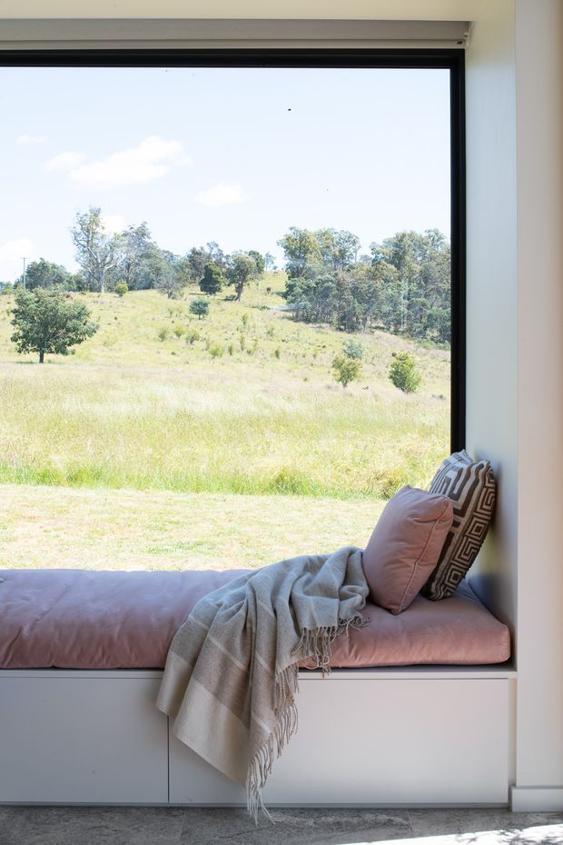 A bright built-in window seat with pale pink cushioning, a grey fringed throw, and patterned cushions. The large black-framed window overlooks a sunny rural landscape, while white built-in cabinetry below and grey flooring create a clean, calm interior setting.