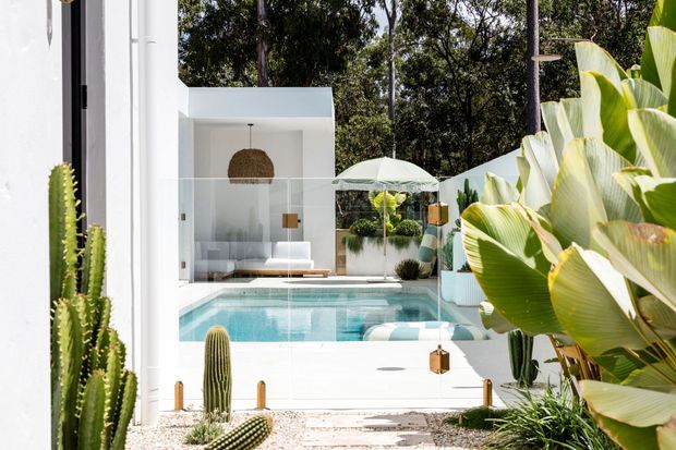Bright contemporary residential pool courtyard viewed from an exterior walkway. A white rendered pavilion with large glazed openings, outdoor seating, pendant lighting, and a turquoise swimming pool is framed by tropical foliage and sculptural cacti. The scene has clean geometric forms, crisp white surfaces, and a relaxed resort-like atmosphere.