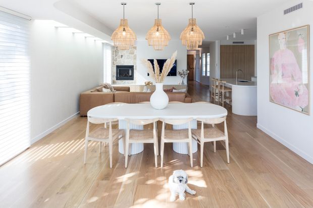 Bright open-plan dining and living area with a light timber floor, white walls, a sculptural white dining table and woven dining chairs. Three woven pendant lights hang above the table, while a tan sectional sofa, fireplace feature wall, artwork and compact kitchen are visible beyond. The room has abundant natural light and a relaxed contemporary coastal feel.