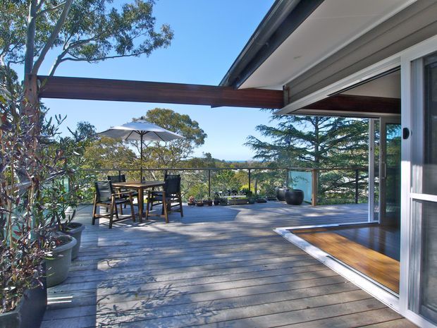 A bright outdoor timber deck and terrace viewed from an open-plan interior, with large sliding glass doors, a timber beam and covered roof overhang. The deck includes an outdoor dining table with chairs, umbrella, potted plants and a glass balustrade, overlooking a leafy landscape and distant blue horizon.