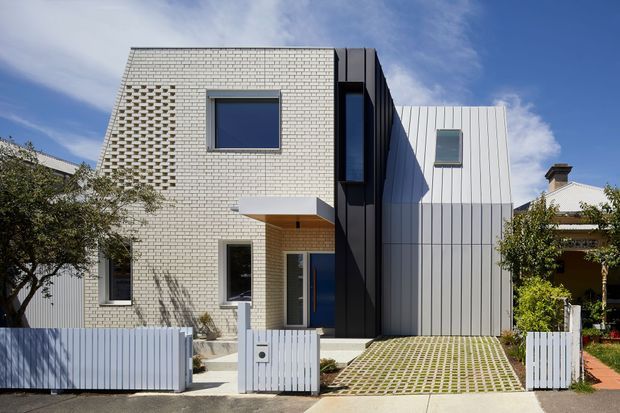 Street-facing exterior of a contemporary multi-level residence with a pale brick upper volume, dark vertical central cladding, and a light grey corrugated metal-clad side volume. The facade includes varied rectangular windows, a recessed entry beneath a timber-toned soffit, a white picket-style fence and gate, and a permeable grid driveway. A neighboring traditional home is partially visible to the side.