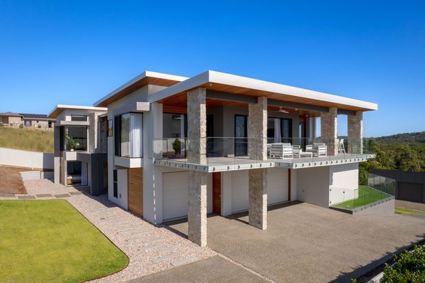 Exterior architectural photograph of a contemporary two-storey residence with a flat roof, projecting upper-level terrace, extensive glazing, white rendered walls, dark framed windows, timber soffit accents, and prominent rough stone-clad columns. The home sits on a landscaped sloping site with a broad driveway and lawn.